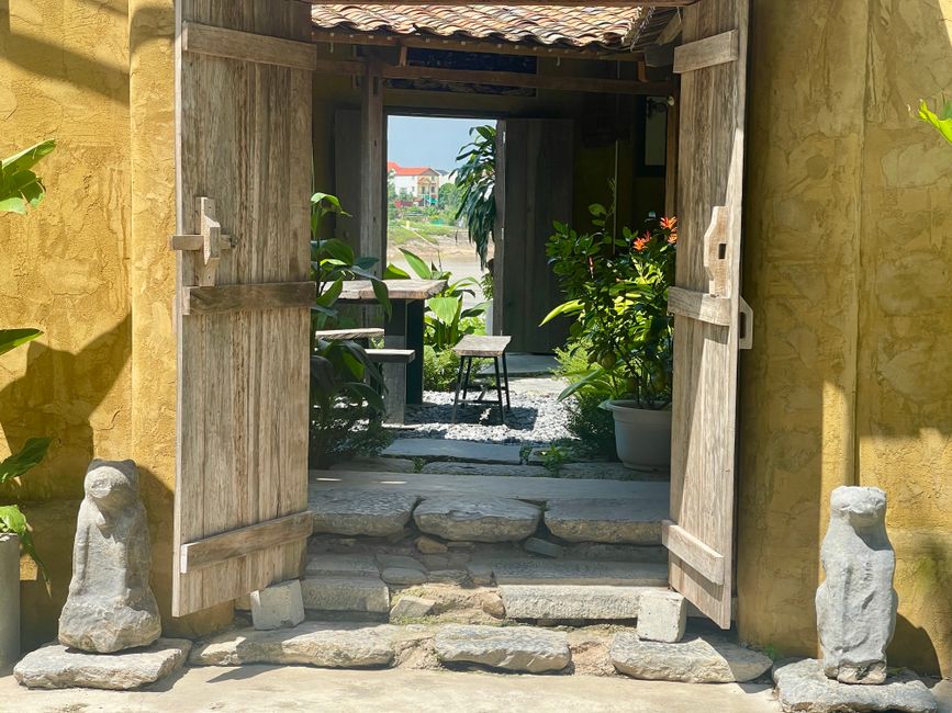 Charming wooden gate opens to a sunlit garden with stone statues, evoking tranquility.